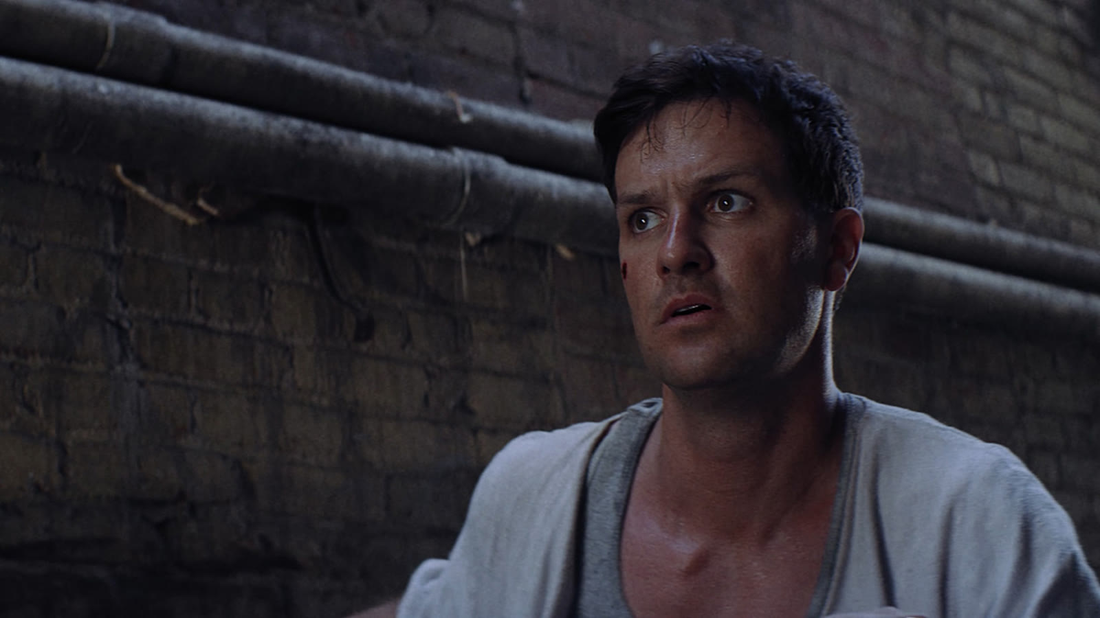 A man pressed against a brick wall, looking off-frame in alarm.